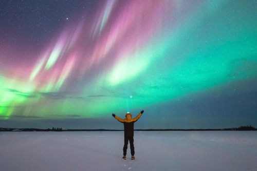 A person is standing in the snow with their arms outstretched in front of the aurora borealis