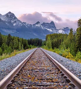 Sunset view of railroad tracks stretching towards the snow-capped Canadian Rockies, surrounded by lush green pine trees.